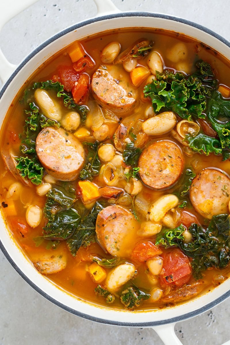 A pot of Italian sausage and white bean soup with kale sitting on a stone surface.