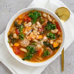 Italian sausage and kale soup with white beans in a bowl on a napkin with a spoon sitting next to it.